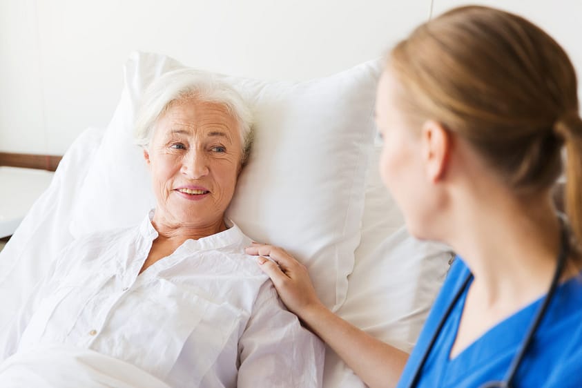 medicine, age, support, health care and people concept - doctor or nurse visiting and cheering senior woman lying in bed at hospital ward
