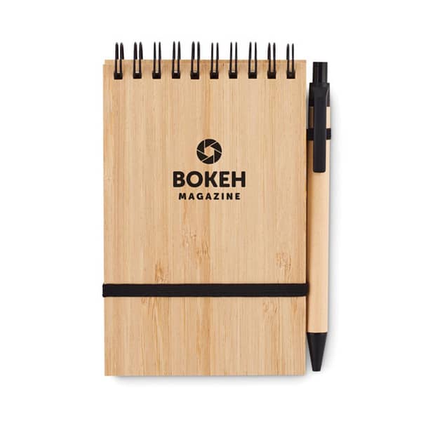 Notebook with logo SONORABAM A6 bamboo notepad with logo and hard cover. Twin-wire. 140 plain recycled paper pages (70 sheets). Matching push button ball pen with recycled paper barrel and PLA detailing. Blue ink. Elastic closure strap and pen holder. Depending on the surface we can use embroidery, engraving, 360° imprint or screen print.